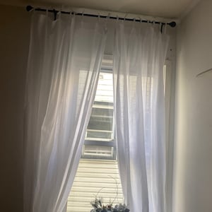 Può includere: Tende bianche e trasparenti sono appese a una finestra con un'asta per tende nera. Una pianta in vaso con foglie verdi e fiori bianchi si trova sul davanzale della finestra.