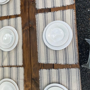 May include: Four white dinner plates with a decorative rim are set on four beige placemats with blue stripes. The placemats have a frayed edge and are laid out on a wooden table.