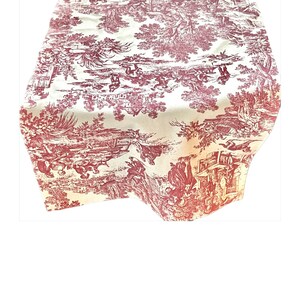 May include: A red and white toile table runner with a repeating pattern of trees, houses, and figures. The runner is made of a soft, woven fabric.