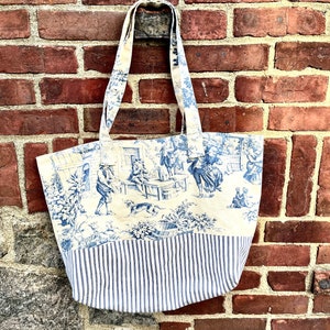Può includere: Una borsa tote bianca con tessuto a righe blu e bianche nella parte inferiore. La parte superiore della borsa presenta un motivo toile blu e bianco con una scena di persone e animali.