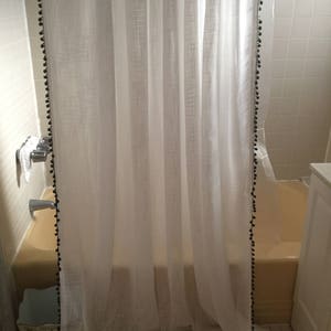 Può includere: Una tenda da doccia bianca con un bordo di pon pon neri è appesa in un bagno. La tenda da doccia è realizzata in un tessuto trasparente e ha un motivo testurizzato. Il bagno ha pareti piastrellate bianche e una vasca bianca.