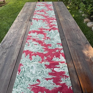 May include: A red and white toile table runner with a scenic design of trees, buildings, and figures. The runner is placed on a weathered wooden table, adding a touch of elegance to the rustic setting.