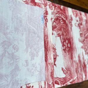 Pu&ograve; includere: Un runner da tavolo rosso e bianco con un motivo toile che rappresenta una scena di caccia. Il runner ha un pannello centrale in lino bianco e un bordo rosso e bianco.