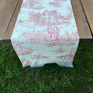 Può includere: Un runner da tavolo bianco con un motivo toile rosso e bianco che presenta un cottage, alberi e persone. Il runner è su un tavolo di legno e lo sfondo è erba verde.
