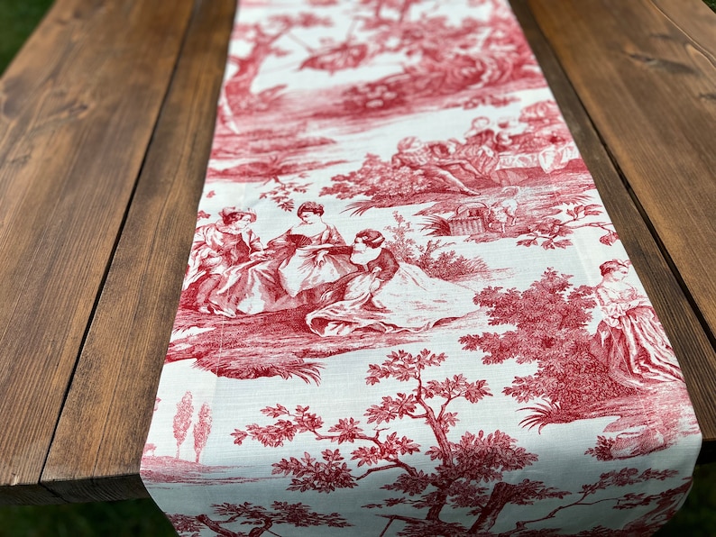 Pu&ograve; includere: Tovaglia da tavolo in toile rossa e bianca con un design country francese che presenta una scena pastorale con persone e alberi.