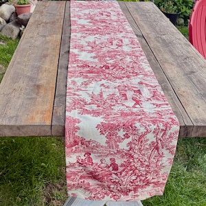 Può includere: Una tovaglia in toile de Jouy rossa e bianca con un disegno di scena pastorale. La tovaglia è drappeggiata su un tavolo di legno, mostrando un motivo dettagliato di figure, edifici e alberi. Il tessuto sembra essere cotone o lino.