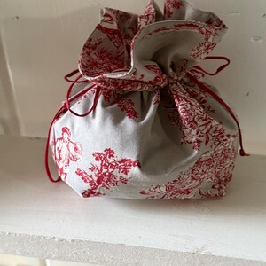 May include: A gray drawstring bag with a red toile pattern featuring figures and floral designs. The bag is cinched closed with a red cord.