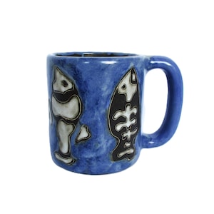 May include: A blue ceramic mug with a mottled finish featuring two dark, stylized fish skeleton illustrations on the exterior. The mug has a thick, rounded handle and a cylindrical body with a slightly uneven, hand-crafted texture throughout.
