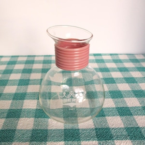 May include: A clear glass carafe with a round base and a pink, ribbed collar. The top is flared and angled. The carafe has a logo that says "MERS" and "CORY" printed on the front. The carafe is sitting on a green and white checkered tablecloth.