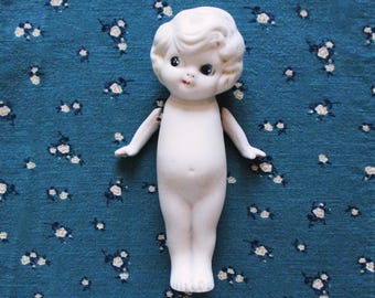 May include: A small, antique bisque doll with a white body and curly hair. The doll has painted facial features, including blue eyes and a small red mouth. The doll is standing on a blue floral fabric.