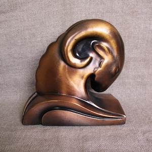 May include: A bronze-colored sculpture of a ram's head with curved horns. The sculpture has a smooth, polished finish and is mounted on a rectangular base. The ram's head is detailed with a focus on the horns and facial features.