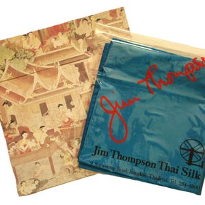 May include: Two fabric pouches. One features a detailed, multi-colored illustration of a traditional Thai scene. The other is a teal blue bag with the red text "Jim Thompson" and "Jim Thompson Thai Silk" printed on it.