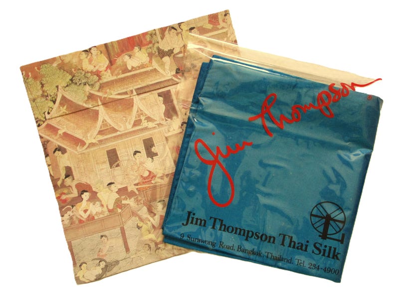May include: Two fabric pouches. One features a detailed, multi-colored illustration of a traditional Thai scene. The other is a teal blue bag with the red text "Jim Thompson" and "Jim Thompson Thai Silk" printed on it.