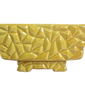 Puede incluir: Una maceta rectangular de cerámica con un acabado amarillo mostaza brillante. La superficie exterior presenta una textura geométrica cristalina y facetada compuesta por numerosas formas triangulares y poligonales pequeñas.