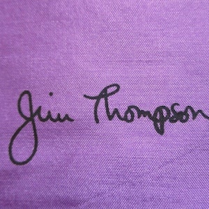 May include: A solid purple fabric surface featuring the black text Jim Thompson printed in a cursive script style. The material displays a visible woven texture throughout the background, providing a consistent colour and pattern across the entire frame.