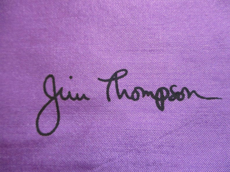 May include: A solid purple fabric surface featuring the black text Jim Thompson printed in a cursive script style. The material displays a visible woven texture throughout the background, providing a consistent colour and pattern across the entire frame.