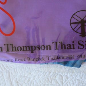 May include: A purple fabric bag featuring a red graphic, a black spinning wheel icon, and the black text Jim Thompson Thai Silk, Surawong Road, Bangkok, Thailand. Tel. 234-4900 printed on the front surface of the smooth, semi-transparent material.