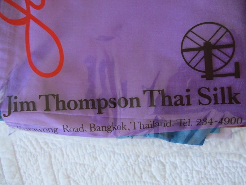 May include: A purple fabric bag featuring a red graphic, a black spinning wheel icon, and the black text Jim Thompson Thai Silk, Surawong Road, Bangkok, Thailand. Tel. 234-4900 printed on the front surface of the smooth, semi-transparent material.
