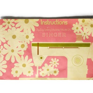 May include: A rectangular instruction manual for a Singer Zig-Zag Sewing Machine Model 242. The cover features a pink background with white and yellow daisy illustrations, a stylized cream-colored sewing machine, and the text Instructions.