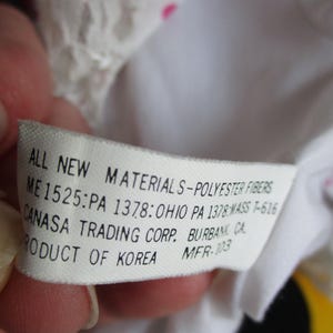 以下が含まれることがあります： 手で持たれた、黒い文字が書かれた白いタグ。タグには「ALL NEW MATERIALS-POLYESTER FIBERS」とその他の詳細が記載されています。タグはCANASA TRADING CORP.のもので、韓国製です。背景は白とピンクです。