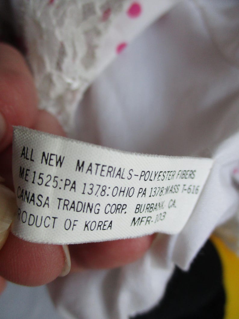 以下が含まれることがあります： 手で持たれた、黒い文字が書かれた白いタグ。タグには「ALL NEW MATERIALS-POLYESTER FIBERS」とその他の詳細が記載されています。タグはCANASA TRADING CORP.のもので、韓国製です。背景は白とピンクです。