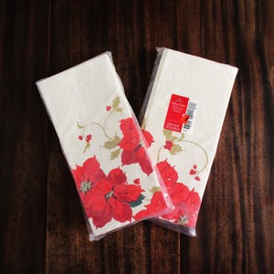 May include: Two packages of cream-colored paper napkins with a red poinsettia floral design. The napkins are packaged in clear plastic and rest on a dark brown wooden surface. The poinsettias have green leaves and red berries.