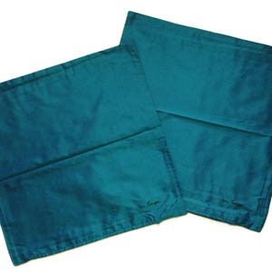 May include: Two teal-colored pillowcases with a subtle sheen. The pillowcases are rectangular and appear to be made of a smooth fabric. Each pillowcase has a small, embroidered signature in the lower corner. The overall aesthetic is simple and elegant.