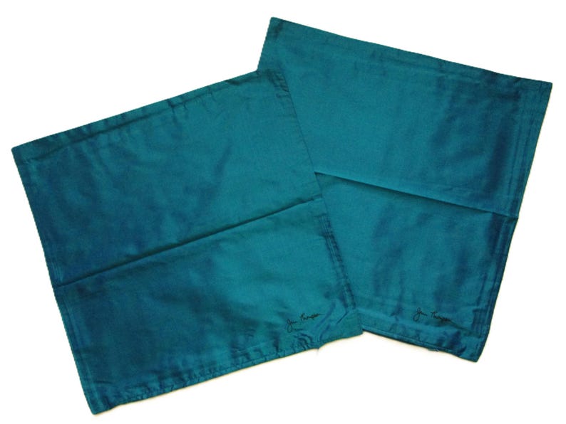 May include: Two teal-colored pillowcases with a subtle sheen. The pillowcases are rectangular and appear to be made of a smooth fabric. Each pillowcase has a small, embroidered signature in the lower corner. The overall aesthetic is simple and elegant.