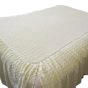 May include: A cream-colored bedspread with a textured, woven pattern. The bedspread has a rectangular top with a ruffled skirt that hangs down around the bed. The fabric appears soft and has a subtle sheen.