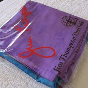 May include: A stack of folded fabric items encased in a clear plastic bag. The top layer is purple with the red text Jim Thompson and a black spinning wheel logo. The text Jim Thompson Thai Silk and an address are printed in black on the right side.