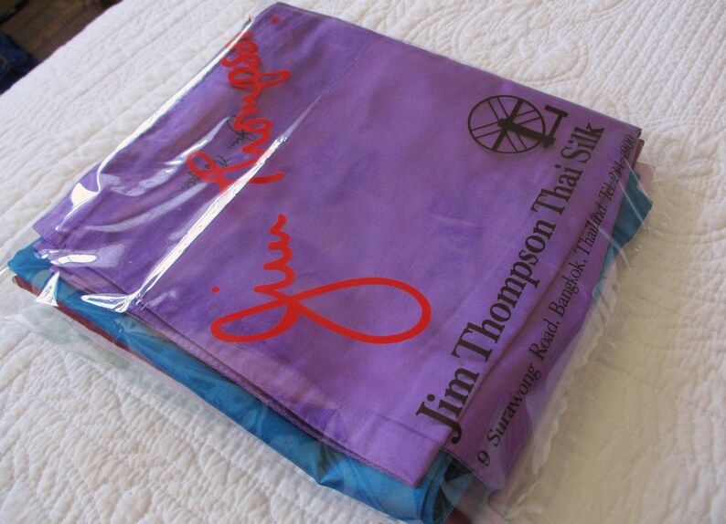 May include: A stack of folded fabric items encased in a clear plastic bag. The top layer is purple with the red text Jim Thompson and a black spinning wheel logo. The text Jim Thompson Thai Silk and an address are printed in black on the right side.