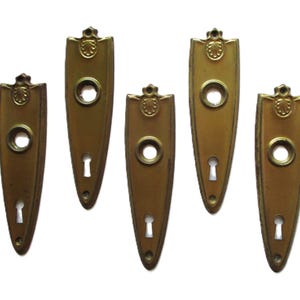以下が含まれることがあります： 5つのアンティーク真鍮製ドアプレート。各プレートは、鍵穴、丸い開口部、装飾的な上部を備えた細長い楕円形です。プレートは暖かく、古びた風合いです。