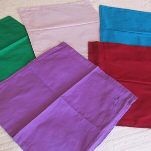 May include: A collection of five rectangular fabric cushion covers in solid colours including green, light pink, blue, purple, and red. The purple cover features a small, dark, handwritten signature in the bottom right corner of the fabric surface.