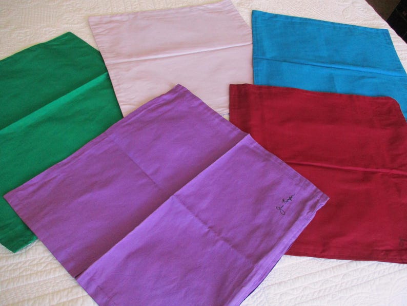 May include: A collection of five rectangular fabric cushion covers in solid colours including green, light pink, blue, purple, and red. The purple cover features a small, dark, handwritten signature in the bottom right corner of the fabric surface.