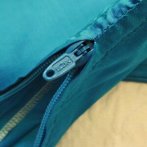 May include: Close-up of a teal-colored zipper with a silver pull tab. The zipper is partially open, revealing the fabric underneath. The zipper pull tab has the word "UCATK" printed on it.