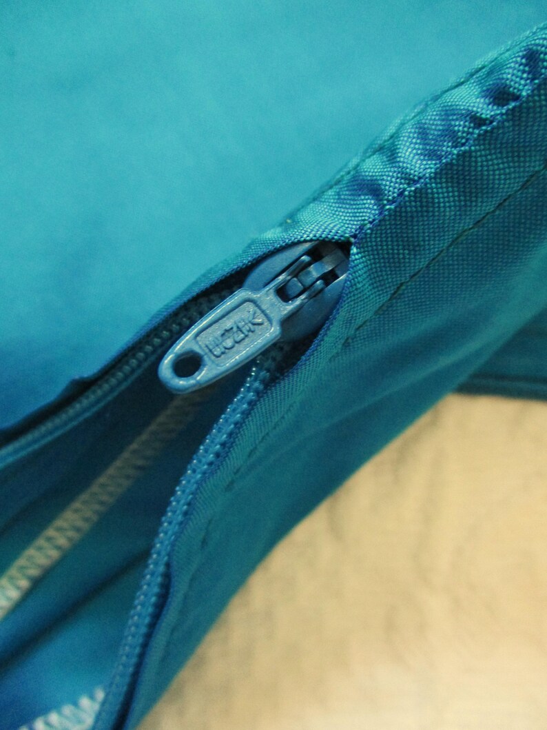 May include: Close-up of a teal-colored zipper with a silver pull tab. The zipper is partially open, revealing the fabric underneath. The zipper pull tab has the word "UCATK" printed on it.
