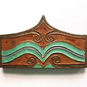 May include: A rectangular ceramic tile with a pointed top edge, featuring a brown textured base with a raised, glossy turquoise wave pattern and symmetrical scroll details. The surface has a matte finish on the brown areas and a smooth glaze.