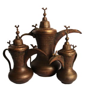 May include: A set of three antique-style copper-toned metal coffee pots, known as dallahs, featuring ornate engraved patterns, curved spouts, and lids topped with small bird-shaped finials. Each vessel has a rounded base and a slender handle.