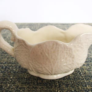 May include: Cream-colored ceramic gravy boat with a handle and a leaf-like texture. The boat has a scalloped rim and a spout for easy pouring. A classic piece for serving sauces and gravies.