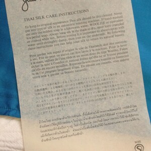 May include: A white paper with black text, featuring care instructions for Thai silk. The paper is from Jim Thompson Thai Silk, with the company's logo and address at the bottom. The paper is on a blue background.