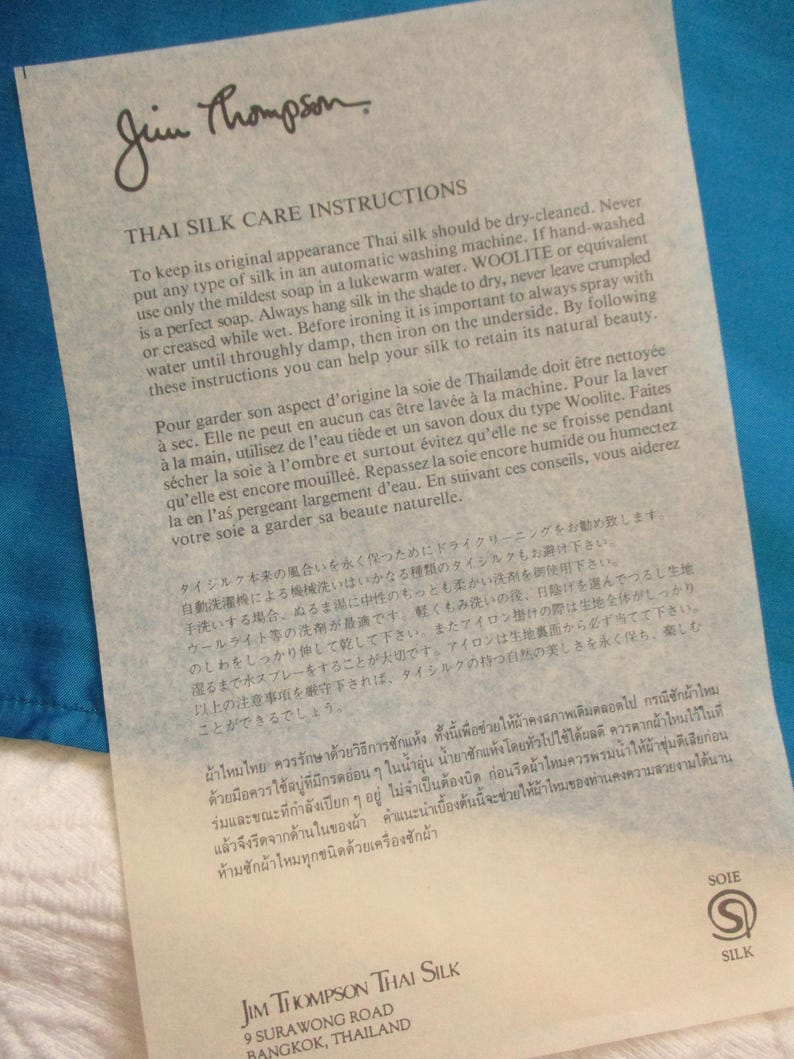 May include: A white paper with black text, featuring care instructions for Thai silk. The paper is from Jim Thompson Thai Silk, with the company's logo and address at the bottom. The paper is on a blue background.