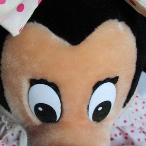 以下が含まれることがあります： 桃色の顔、大きな白い目と黒い瞳、黒い鼻をしたキャラクターのぬいぐるみ。おもちゃは頭の上に黒い毛皮があり、ピンクのドットが付いた白いボンネットをかぶっています。