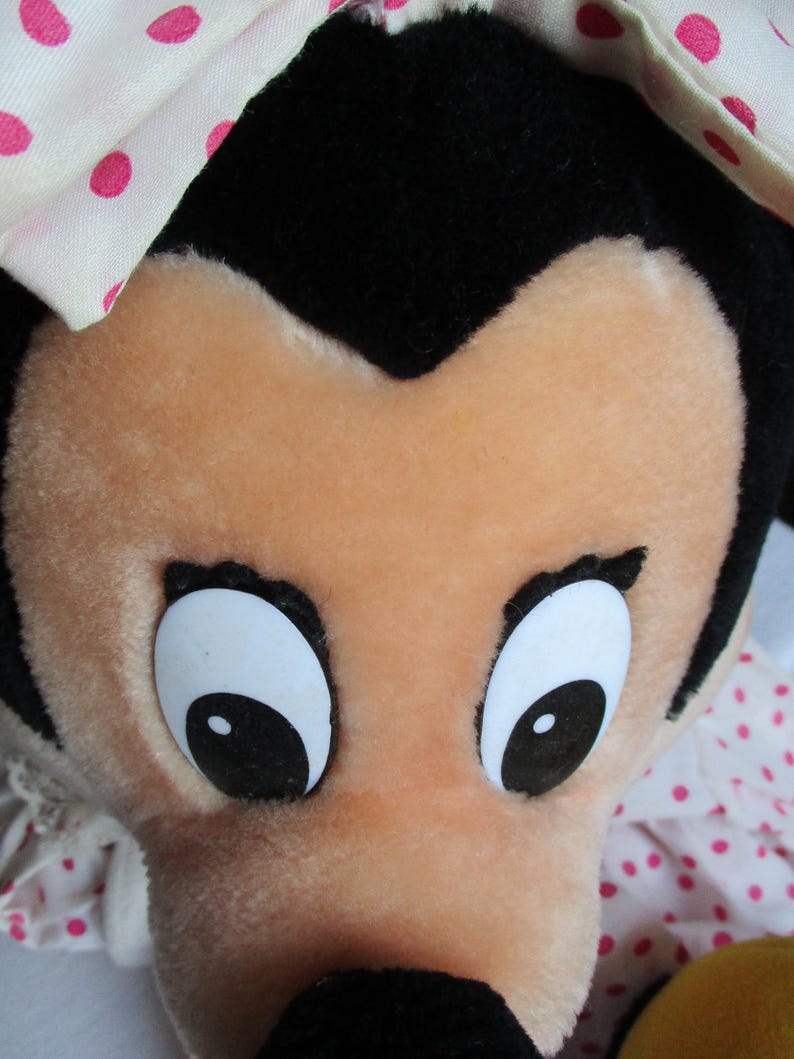 以下が含まれることがあります： 桃色の顔、大きな白い目と黒い瞳、黒い鼻をしたキャラクターのぬいぐるみ。おもちゃは頭の上に黒い毛皮があり、ピンクのドットが付いた白いボンネットをかぶっています。
