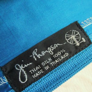 May include: A bright blue fabric item featuring a black rectangular label with white text that reads Jim Thompson, THAI SILK 100%, MADE IN THAILAND, accompanied by a small graphic of a spinning wheel icon stitched onto the material.