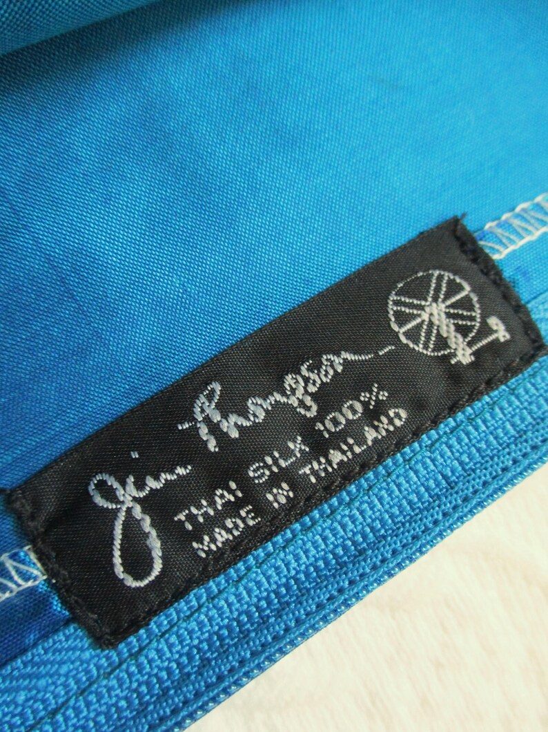 May include: A bright blue fabric item featuring a black rectangular label with white text that reads Jim Thompson, THAI SILK 100%, MADE IN THAILAND, accompanied by a small graphic of a spinning wheel icon stitched onto the material.