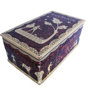 May include: A rectangular box with a dark purple and black marbled pattern, featuring cream-colored illustrations of figures and trees. The lid displays the word GUERLAIN in cream lettering, surrounded by a decorative border of leaves and vines.