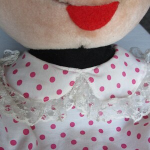 以下が含まれることがあります： ぬいぐるみ人形の顔と服のクローズアップ。人形は薄いピンク色の顔、赤いフェルトの笑顔、黒い襟をしています。白いドレスにはピンクのドットとレーストリムの襟が飾られています。