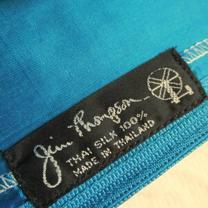 May include: Close-up of a vibrant turquoise fabric with a textured weave. A black label with white script reads "Jim Thompson" and "Thai Silk 100% Made in Thailand". The label features a logo of a stylized wheel.