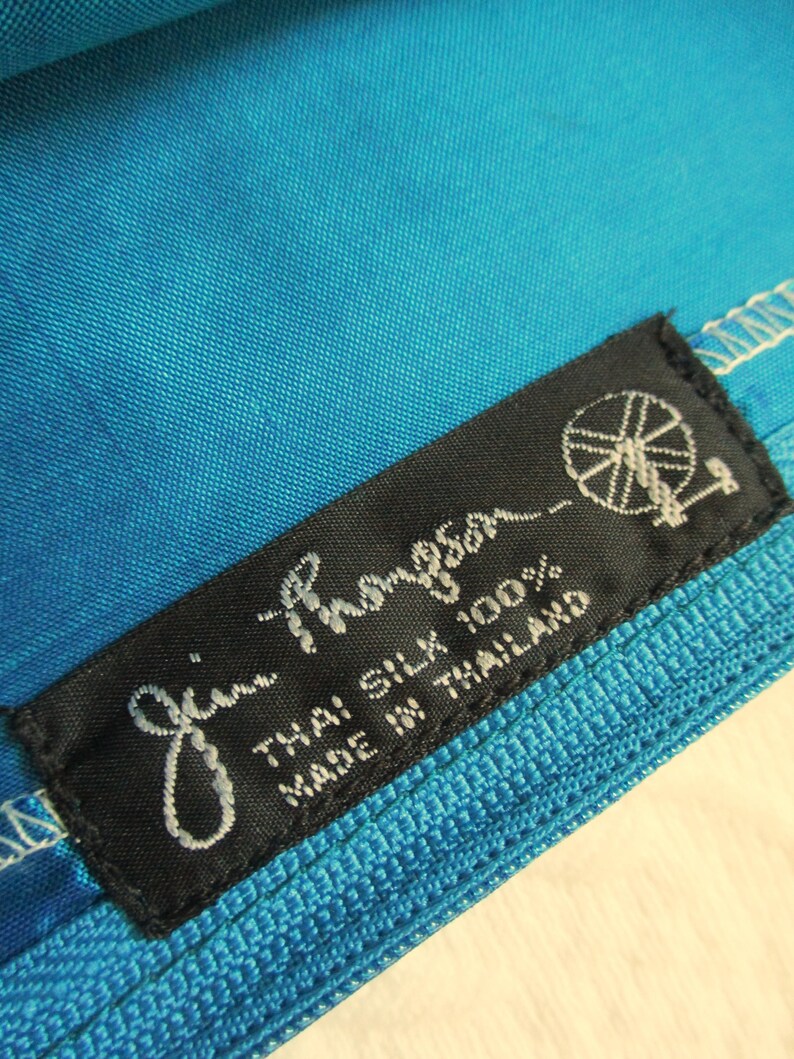 May include: Close-up of a vibrant turquoise fabric with a textured weave. A black label with white script reads "Jim Thompson" and "Thai Silk 100% Made in Thailand". The label features a logo of a stylized wheel.
