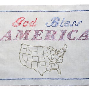 May include: Hand-stitched textile art with the words "God Bless AMERICA" in red, blue, and purple cross-stitch. A map of the United States is embroidered in gold thread below the text. The piece is framed with a blue cross-stitch border.